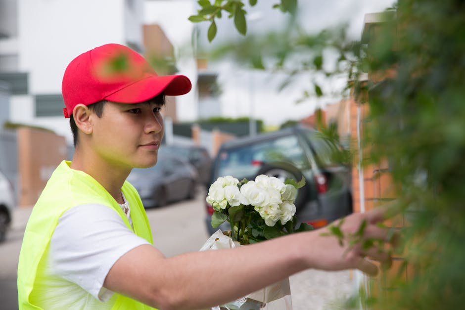 A young male florist wearing a red cap and a high-visibility yellow vest is arranging a bouquet of fresh, white flowers, likely roses or peonies, with lush green foliage. He is outdoors, standing near