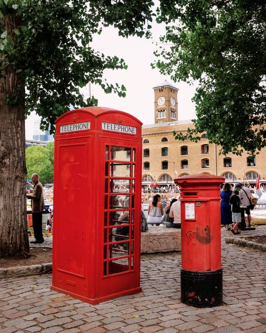 A classic red British telephone box and a smaller red pillar post box are situated outdoors on a cobblestone pavement, surrounded by green foliage from large leafy trees. The telephone box has glass-p