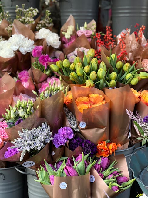 A vibrant display of freshly cut flowers arranged in brown paper wrapping, featuring a mix of tulips, carnations, and other seasonal blooms. The tulips are predominantly pink and purple, with some sti
