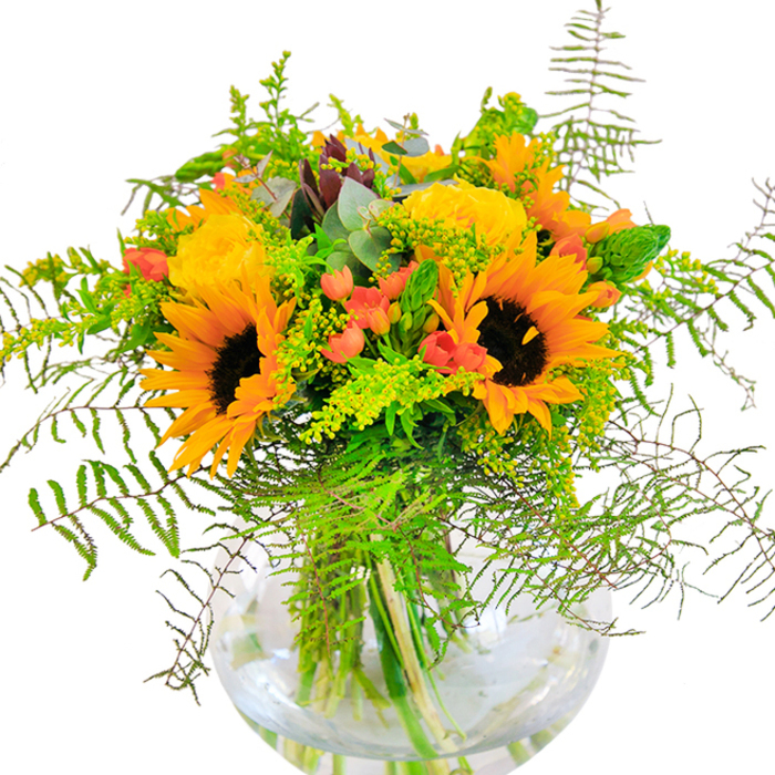 First impressions of this Kings Cross bouquet are shaped by an exuberant glow-sunflowers and buttery yellow garden roses radiate warmth and promise, their open petals arranged in generous, embracing layers. Scarlet hypericum berries, small and lustrous, punctuate the design with lively, celebratory red against deep green leaves and lighter cascades of airy fern. The composition weaves in sprigs of solidago and slender flower spikes, echoing the motion and energy of travelers at St Pancras. Bluish-green eucalyptus introduces a cooling undertone, its rounded leaves enhancing the natural, rustic elegance. Everything is artfully presented in a spherical glass vase, whose transparent curves reveal clean lines of water-bathed stems, reflecting the contemporary style of nearby York Way. The bouquet's textures contrast and complement-from the velvety sunflower heads to the silky rose petals and crisp freshness of new-growth foliage. Its subtle fragrance carries hints of summer meadows and freshly cut greens, inviting one to linger and savor the bouquet's uplifting presence. Perfect for marking birthdays, milestones, or simply sharing a moment of joy with friends along Pancras Road, this bouquet is an emblem of Kings Cross's welcoming, creative heart.